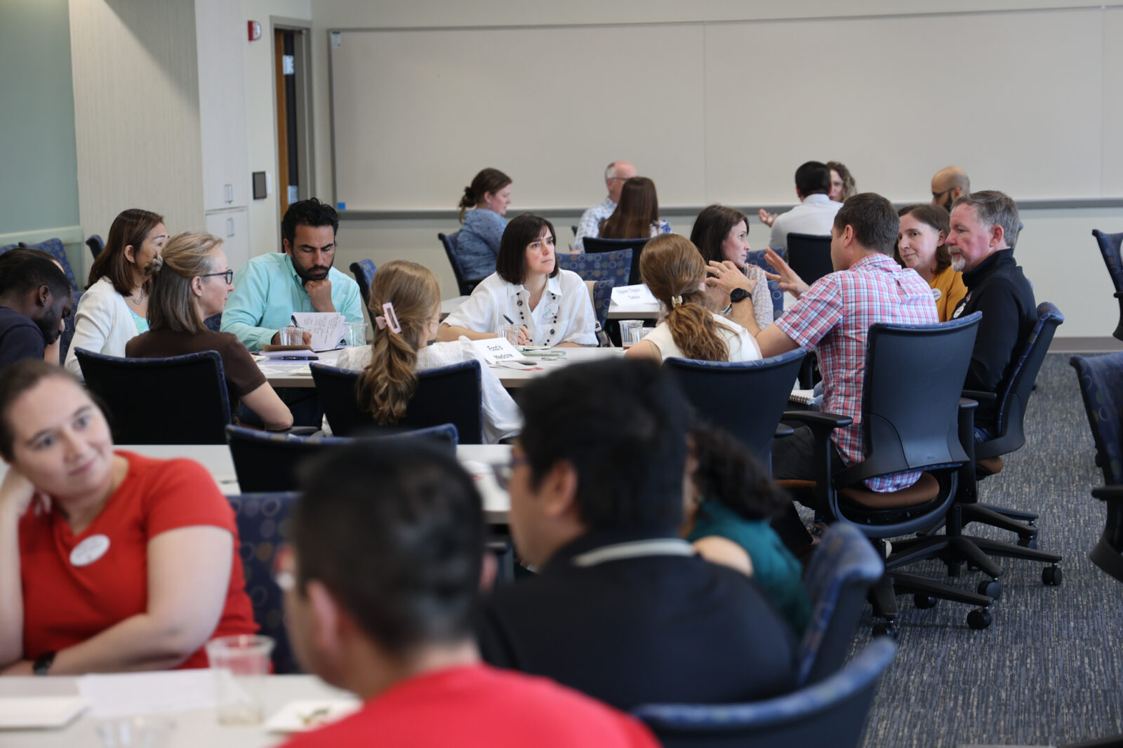Researchers sit in small groups around tables, talking and taking notes during a sustainability research networking session.
