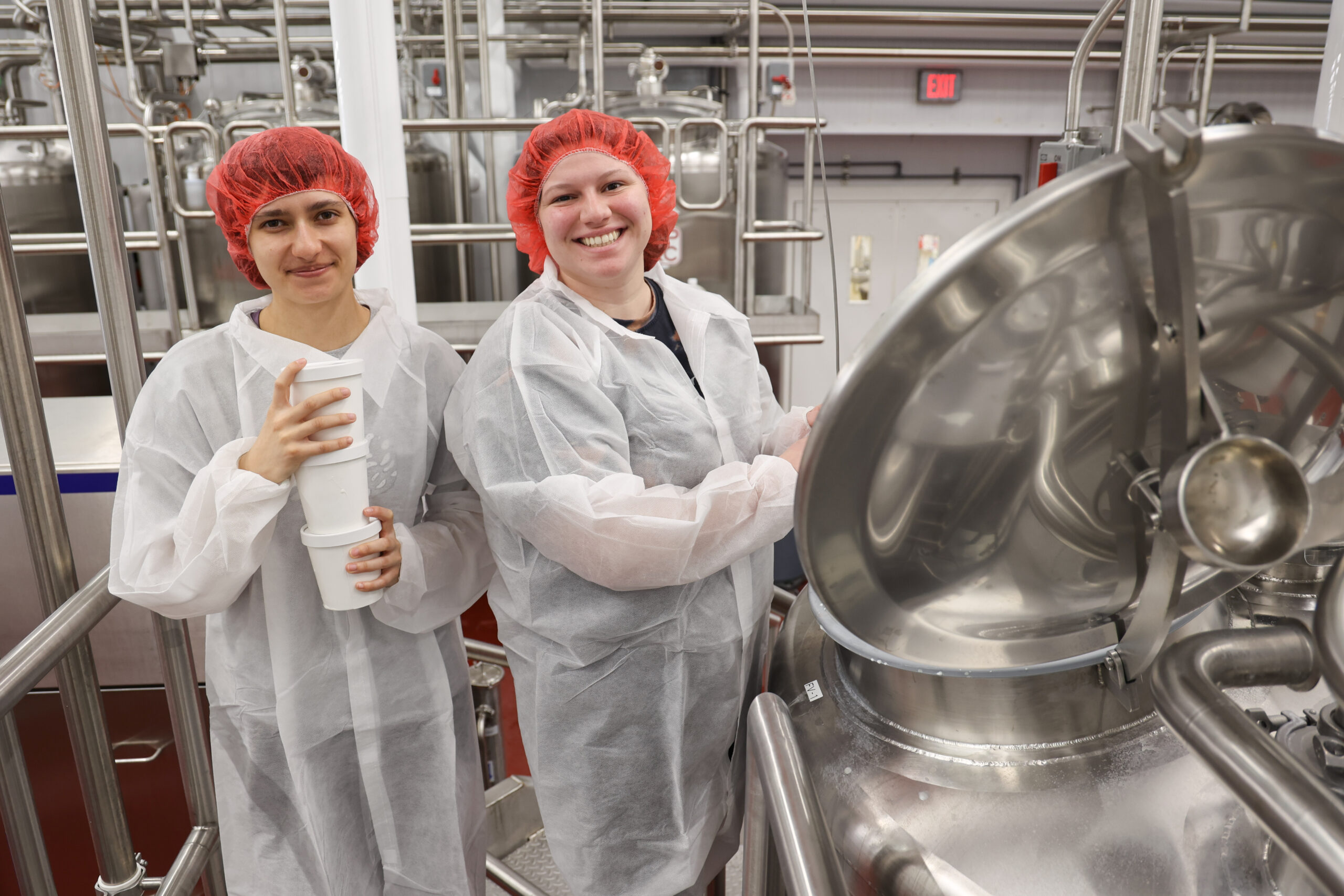 Loren Smith and Sydney Camp smile beside ice cream production equipment at Babcock Dairy, wearing white protective gowns and red hairnets.