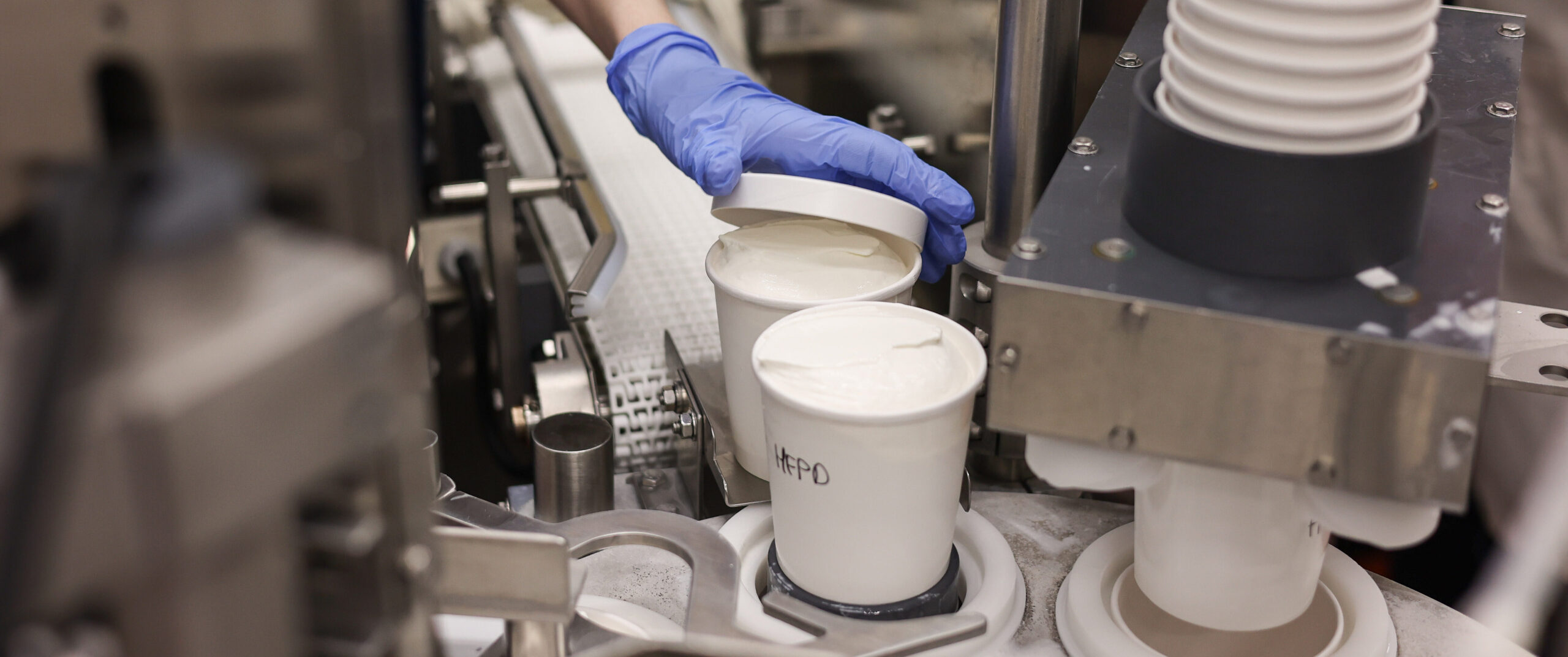 A blue-gloved hand places a lid on a pint of vanilla ice cream as filled containers move through packaging equipment.