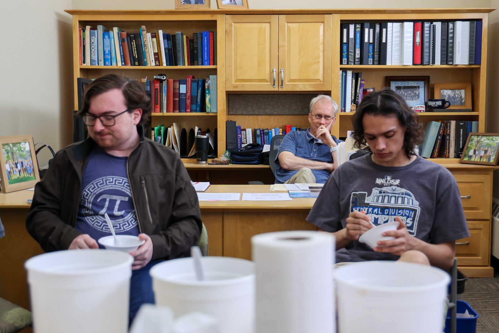 Three people seated in an office taste ice cream from bowls, with sample containers on the table in the foreground.