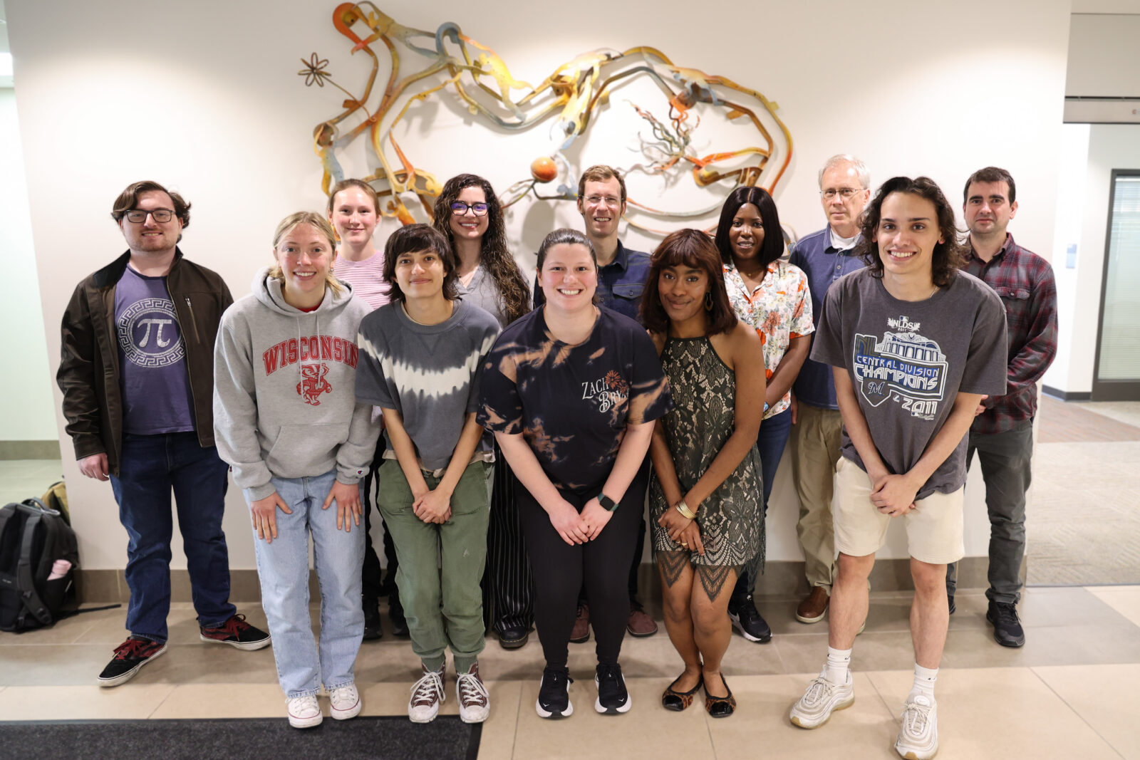 Twelve project team members pose for a group photo in two rows, standing in front of a colorful abstract wall sculpture.